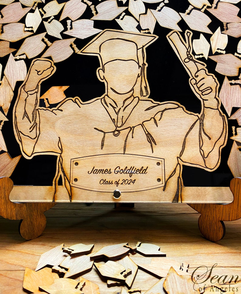 K&ouml;nnte beinhalten: Holzhalter f&uuml;r Abschlusskappen mit einer Aussparung eines Absolventen, der ein Diplom h&auml;lt. Der Halter hat eine personalisierte Namensschilder mit der Aufschrift "James Goldfield Klasse von 2024".