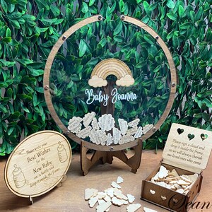 May include: A wooden baby shower guest book with a clear circular frame, a rainbow, and the name "Baby Joanna." Includes a sign, wooden clouds, and a box for guests to sign and drop in the frame. The sign says "Please write your Best Wishes for the New Baby."
