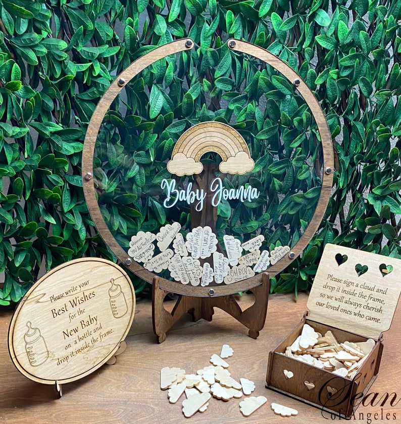 May include: A wooden baby shower guest book with a clear circular frame, a rainbow, and the name "Baby Joanna." Includes a sign, wooden clouds, and a box for guests to sign and drop in the frame. The sign says "Please write your Best Wishes for the New Baby."