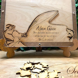 May include: A wooden plaque with a laser-cut design of a fisherman casting a line with a fish jumping out of the water. The plaque is personalized with the name "Adam Cohen" and the text "Your dedication and perseverance set the bar for the rest of us. You'll be missed."