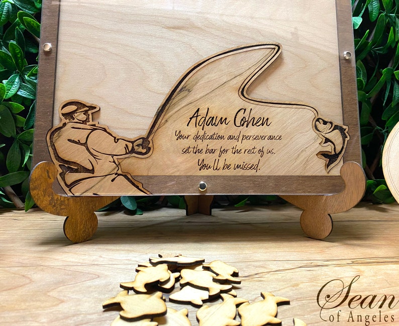 May include: A wooden plaque with a laser-cut design of a fisherman casting a line with a fish jumping out of the water. The plaque is personalized with the name "Adam Cohen" and the text "Your dedication and perseverance set the bar for the rest of us. You'll be missed."