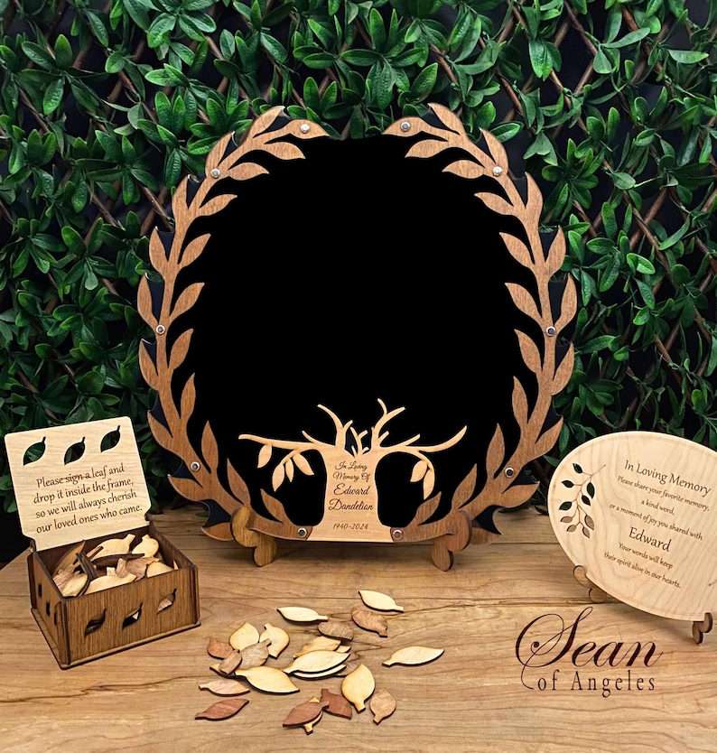 May include: A wooden memorial tree frame with a black background and a tree with leaves. The frame is surrounded by a wreath of leaves. There is a small wooden box with leaves inside and a sign that says "Please sign a leaf and drop it inside the frame, so we will always cherish our loved ones who came." There is also a small wooden plaque with the text "In Loving Memory of Edward Dandelion 1940-2024".