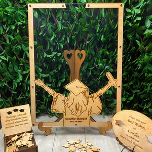 Op de afbeelding: Houten herinneringsdoos voor de afstuderen met een helder acryl frame, een houten uitsnijding van een afgestudeerde en een klein houten doosje gevuld met houten afstudeerhoedjes. Het frame heeft de tekst "Onderteken een chip en laat hem in het frame vallen, zodat we onze dierbaren die zijn gekomen altijd zullen koesteren". De uitsnijding heeft de tekst "Camilla Claudel Klas van 2023". Het kleine houten doosje heeft de tekst "Sean of Angeles".