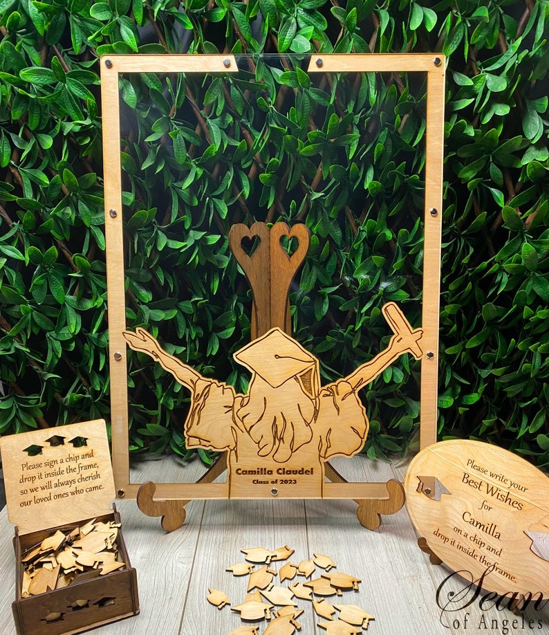 Op de afbeelding: Houten herinneringsdoos voor de afstuderen met een helder acryl frame, een houten uitsnijding van een afgestudeerde en een klein houten doosje gevuld met houten afstudeerhoedjes. Het frame heeft de tekst "Onderteken een chip en laat hem in het frame vallen, zodat we onze dierbaren die zijn gekomen altijd zullen koesteren". De uitsnijding heeft de tekst "Camilla Claudel Klas van 2023". Het kleine houten doosje heeft de tekst "Sean of Angeles".