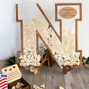 May include: A wooden letter N with a clear acrylic frame filled with small wooden hearts. The letter is standing on a wooden base. A small wooden box with a heart design is in the foreground. The box is filled with wooden hearts. The text "Stephanie & Colton December 4, 2021" is on a small wooden plaque attached to the letter.