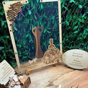 May include: A wooden quincea&ntilde;era guest book with a clear acrylic front. The frame features laser-cut floral and dress designs. Includes a box of wooden hearts and a sign that reads "Please write your Best Wishes for Morgan".