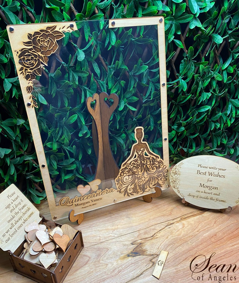 May include: A wooden quincea&ntilde;era guest book with a clear acrylic front. The frame features laser-cut floral and dress designs. Includes a box of wooden hearts and a sign that reads "Please write your Best Wishes for Morgan".