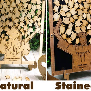 Op de afbeelding: Twee herinneringsdozen voor afstudeerhoeden, een met een natuurlijke houten afwerking en de andere met een gebeitst houten afwerking. Beide dozen hebben een uitgesneden ontwerp van een afgestudeerde die een stapel afstudeerhoeden omhoog houdt. De dozen hebben een transparante acrylglasfront en een houten ezelstandaard. De tekst op de dozen luidt "Camilla Claudel Klas van 2023" en "Yamik Noah Klas van 2023".