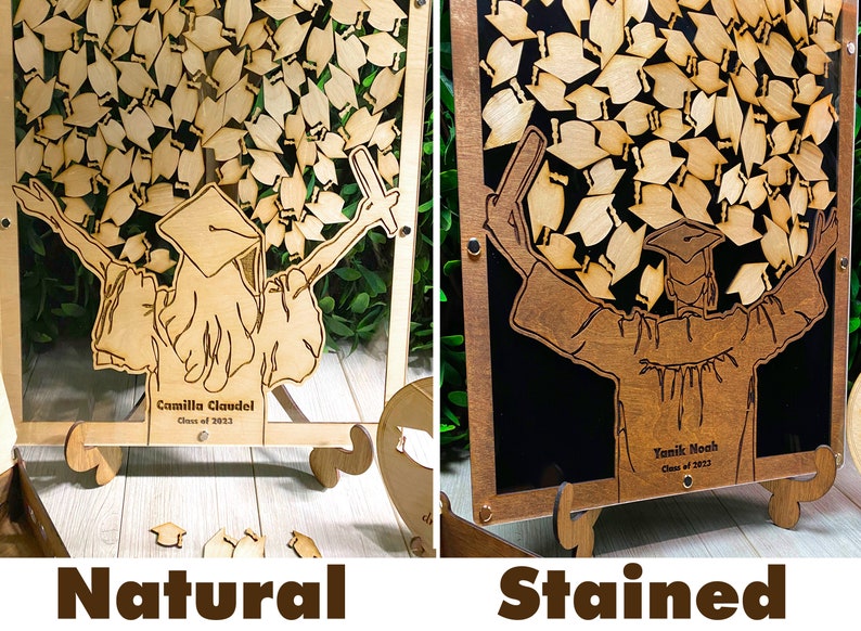 Op de afbeelding: Twee herinneringsdozen voor afstudeerhoeden, een met een natuurlijke houten afwerking en de andere met een gebeitst houten afwerking. Beide dozen hebben een uitgesneden ontwerp van een afgestudeerde die een stapel afstudeerhoeden omhoog houdt. De dozen hebben een transparante acrylglasfront en een houten ezelstandaard. De tekst op de dozen luidt "Camilla Claudel Klas van 2023" en "Yamik Noah Klas van 2023".