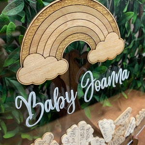 May include: Wooden baby shower keepsake box with a rainbow and cloud design. The words "Baby Joanna" are written in a cursive font. The box is filled with small wooden tags with handwritten messages.
