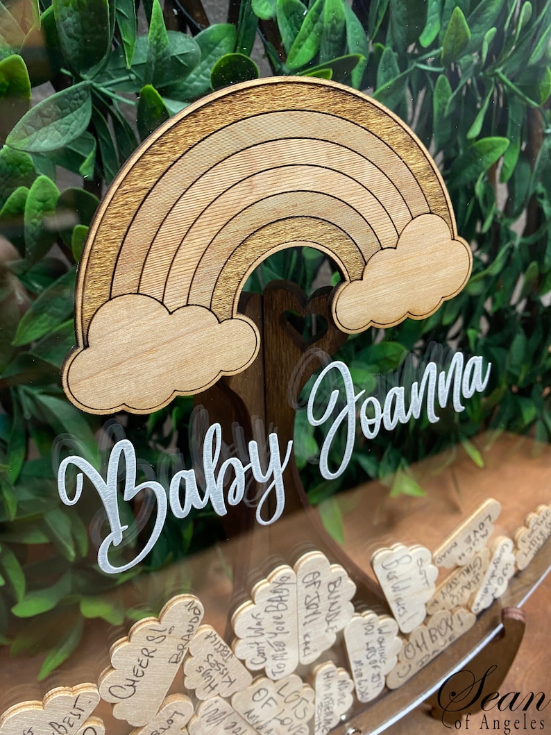May include: Wooden baby shower keepsake box with a rainbow and cloud design. The words "Baby Joanna" are written in a cursive font. The box is filled with small wooden tags with handwritten messages.
