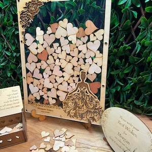 May include: Wooden Quincea&ntilde;era guest book with a frame filled with heart-shaped pieces. The frame has a laser-cut design of a woman in a dress and the text "Quincea&ntilde;era". Includes a wooden box and a sign with text.