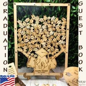 Op de afbeelding: Een houten afstudeer-gastenboek met een transparant acryl frame. Het boek heeft een boom met afstudeerhoeden als bladeren en een afgestudeerde figuur aan de basis. Het frame staat op een houten standaard. De tekst "Graduation" staat aan de linkerkant van het frame en "Guest Book" aan de rechterkant. De tekst "Camilla Claudel Class of 2023" staat onderaan het frame.
