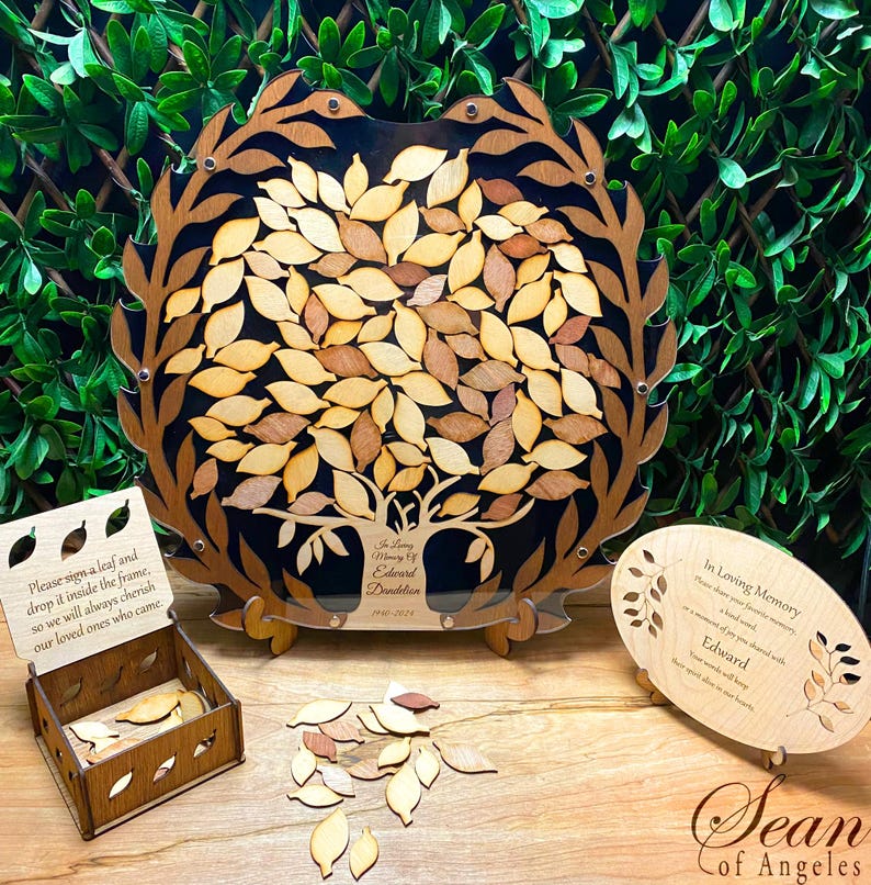 May include: A wooden tree-shaped memorial guest book with a black background and a clear acrylic frame. The tree has many leaves that can be signed by guests. The leaves are made of wood and come in a small wooden box. The guest book also includes a small wooden plaque with the text "In Loving Memory of Edward Dandelion 1941-2023".