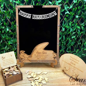 May include: A wooden sign with a chalkboard and a fishing theme. The sign says "Happy Retirement" and "Adam Cohen". There are wooden fish and a small wooden box with a sign that says "Please sign a chip and drop it inside the frame. So we will always cherish our loved ones who came."