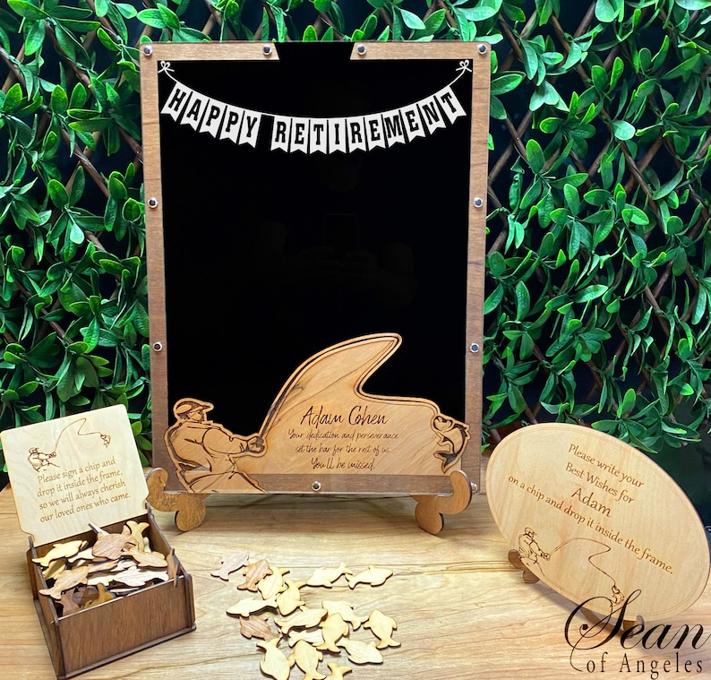 May include: A wooden sign with a chalkboard and a fishing theme. The sign says "Happy Retirement" and "Adam Cohen". There are wooden fish and a small wooden box with a sign that says "Please sign a chip and drop it inside the frame. So we will always cherish our loved ones who came."