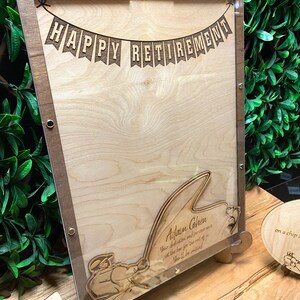 May include: A wooden sign with a clear acrylic overlay, featuring a banner that reads "HAPPY RETIREMENT". The sign also includes a silhouette of a person fishing, with the text "Adam Cohen Your dedication and perseverance at the bar for the rest of... You'll be missed."  The sign is on a wooden stand.