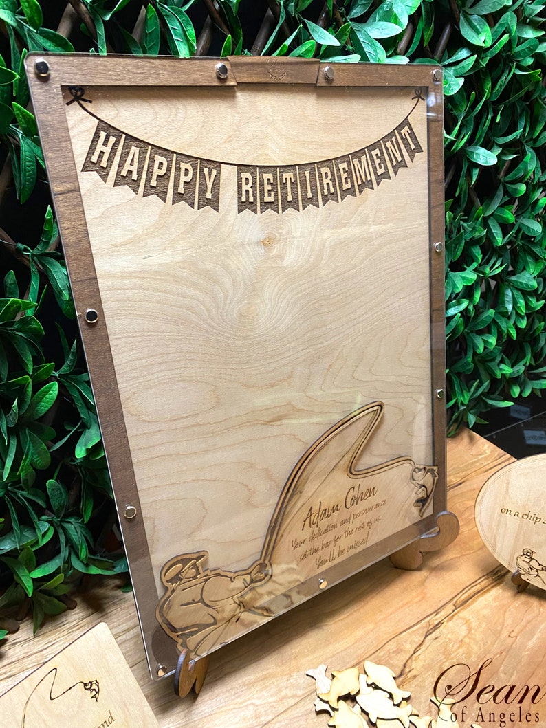 May include: A wooden sign with a clear acrylic overlay, featuring a banner that reads "HAPPY RETIREMENT". The sign also includes a silhouette of a person fishing, with the text "Adam Cohen Your dedication and perseverance at the bar for the rest of... You'll be missed."  The sign is on a wooden stand.