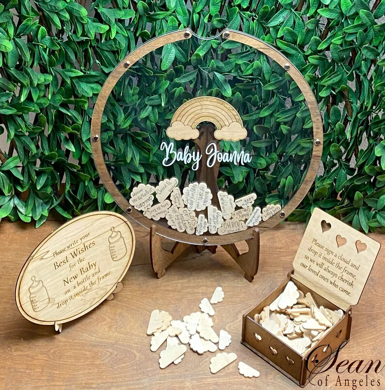 May include: A wooden baby shower guest book with a clear acrylic frame, rainbow, and the name "Baby Joanna." Includes cloud-shaped guest sign-in pieces, a sign with instructions, and a box for the clouds. The frame is supported by a wooden stand.