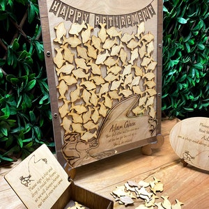 May include: A wooden sign with a fishing theme for a retirement party. The sign has a banner that says "Happy Retirement". There are wooden fish cutouts that guests can sign and drop into a box. The sign also has a personalized message for the retiree, "Adam Cohen, Your dedication and perseverance set the bar for the rest of us. You'll be missed!"