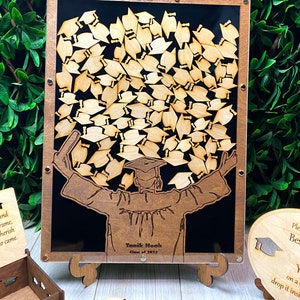 Op de afbeelding: Houten afstudeer-gastenboek met een silhouet van een afgestudeerde die een groot aantal afstudeerhoeden omhoog houdt. Het gastenboek heeft een zwarte achtergrond en een transparante voorkant. De tekst "Yanik Noah Klas van 2023" is gegraveerd op de voorkant van het gastenboek.