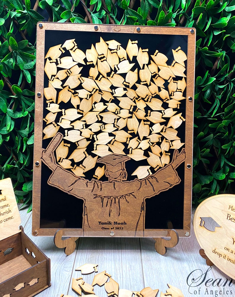 Op de afbeelding: Houten afstudeer-gastenboek met een silhouet van een afgestudeerde die een groot aantal afstudeerhoeden omhoog houdt. Het gastenboek heeft een zwarte achtergrond en een transparante voorkant. De tekst "Yanik Noah Klas van 2023" is gegraveerd op de voorkant van het gastenboek.