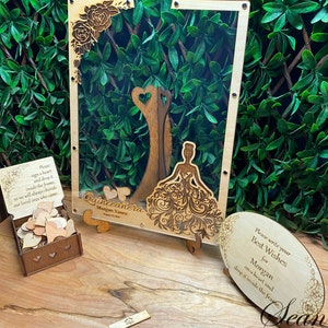 May include: A wooden Quincea&ntilde;era guest book frame with a laser-cut design. The frame includes a wooden box of heart-shaped pieces for guests to sign and drop inside. The frame has the text "Quincea&ntilde;era Morgan Yanez".
