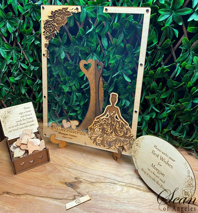May include: A wooden Quincea&ntilde;era guest book frame with a laser-cut design. The frame includes a wooden box of heart-shaped pieces for guests to sign and drop inside. The frame has the text "Quincea&ntilde;era Morgan Yanez".