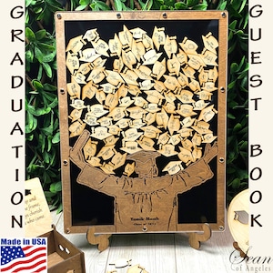 May include: A wooden guest book with a graduation theme. The book features a silhouette of a graduate holding up a diploma with a cap and gown. The background is black and the book is framed in wood. The book is decorated with small wooden graduation caps that guests can sign. The text "GUEST BOOK" is written on the right side of the frame. The text "Yanik Moah Class of 2023" is written on the bottom of the frame.
