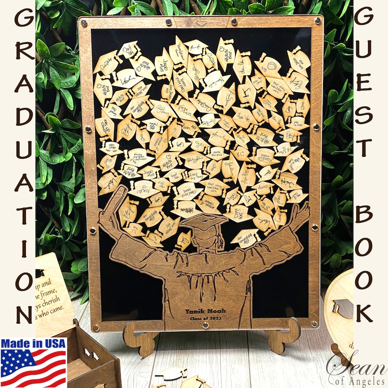 Op de afbeelding: Een houten gastenboek met een afstudeerthema. Het boek toont een silhouet van een afgestudeerde die een diploma vasthoudt met een pet en toga. De achtergrond is zwart en het boek is ingelijst met hout. Het boek is versierd met kleine houten afstudeerpetten die gasten kunnen signeren. De tekst "GUEST BOOK" staat aan de rechterkant van de lijst. De tekst "Yanik Moah Class of 2023" staat onderaan de lijst.