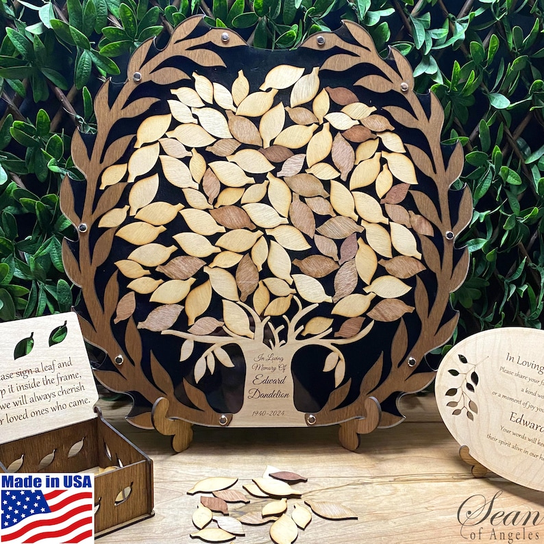 May include: A wooden memorial tree with leaves that can be signed by guests. The tree is on a black background with the words "In Loving Memory of Edward Dandelion 1940-2024". There are extra leaves in a pile below the tree. The words "Made in USA" are on a small wooden box with an American flag.