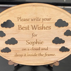 May include: Wooden oval sign with the text "Please write your Best Wishes for Sophie on a cloud and drop it inside the frame." Features eight black cloud cutouts. The sign is supported by a wooden stand.