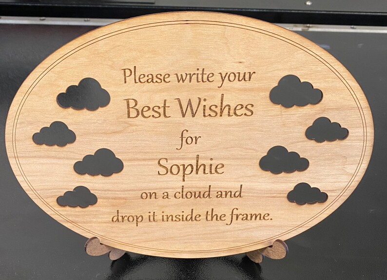 May include: Wooden oval sign with the text "Please write your Best Wishes for Sophie on a cloud and drop it inside the frame." Features eight black cloud cutouts. The sign is supported by a wooden stand.