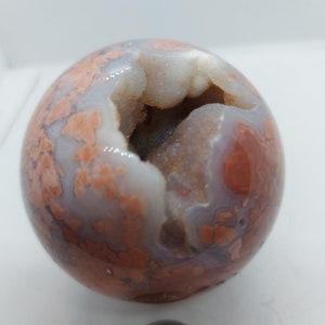 Puede incluir: Una geoda esférica con un centro hueco. El exterior presenta una mezcla de colores, incluyendo blanco, naranja y gris. El interior revela una formación cristalina brillante. Este artículo decorativo es una pieza única de arte natural.