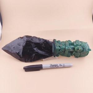 May include: A handcrafted obsidian blade with a detailed green handle featuring carved figures. A black Sharpie marker is placed next to the blade for size comparison. The blade is approximately 10 inches long.