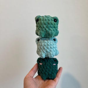May include: Three stacked, handmade crochet frog-shaped items in varying shades of green and blue. Each frog has black button eyes and small pink cheeks. The frogs are held in a hand against a plain white background.