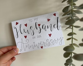 May include: A white card with the words "TO MY Husband ON OUR Anniversary" in black script, accented with small red hearts. The card is held by a hand.