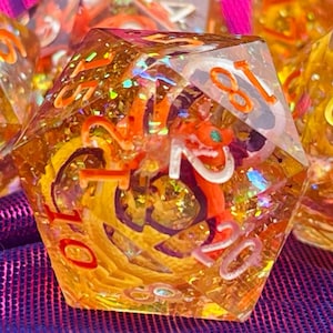 Puede incluir: Primer plano de un juego de seis dados poliédricos. Los dados son transparentes con un tono ámbar, llenos de remolinos y purpurina naranja y dorada. Cada dado tiene números blancos. Los dados están sobre una tela morada y rosa.