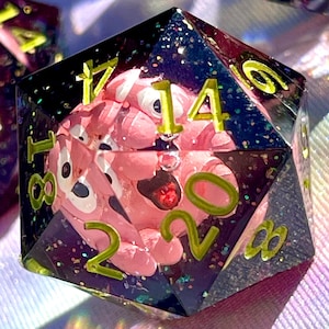 May include: Close-up of several polyhedral dice. The dice are clear with a dark, glittery interior. Each die has a pink, cartoon-like figure inside. The numbers are in a bright green color.