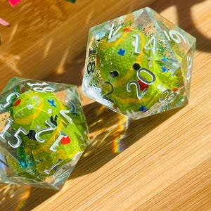 May include: Two clear, multi-sided dice with green and yellow interiors. The dice have white numbers and are filled with glitter and small colorful accents. The dice are set on a wooden surface.