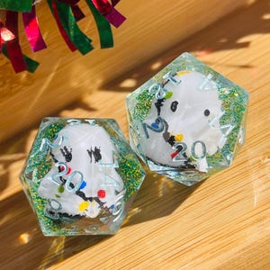 May include: Two polyhedral dice with a snowman design. The dice are clear with a glittery green interior and white snowman figures. The numbers are in black and the snowman has colorful accents. The dice are on a wooden surface.