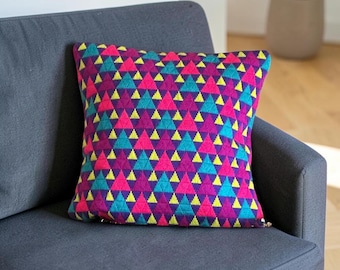 Puede incluir: Un cojín cuadrado con un patrón geométrico repetitivo de pequeños triángulos en tonos vibrantes de rosa intenso, turquesa brillante, amarillo lima y morado oscuro. La superficie textil tiene una apariencia tejida y texturizada.