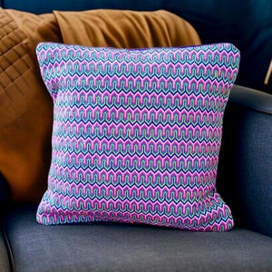 May include: Square decorative pillow with a geometric pattern in shades of pink, blue, and green. The pillow is placed on a gray surface, with other pillows in the background. The pillow has a textured appearance.