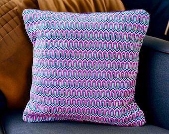 Puede incluir: Un cojín cuadrado con un patrón geométrico repetitivo de chevrón en tonos vibrantes de magenta, azul brillante y verde lima. La tela tiene una apariencia tejida y texturizada con líneas en zigzag verticales constantes en toda la superficie.