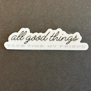 May include: A white sticker with the words "all good things" in a cursive font, and "TAKE TIME. MY FRIEND" in a sans-serif font. The sticker has a cloud-like shape and is on a dark gray background.