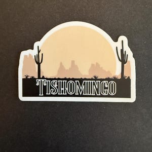 May include: A sticker featuring a desert landscape with a setting sun. Two cacti flank the scene, with the word "TISHOMINGO" in white lettering at the bottom. The background is a gradient of tan and brown.