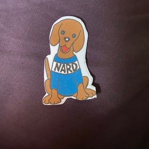 May include: A cartoon illustration of a brown dog wearing a blue shirt with the word "NARD" printed in white on the front.
