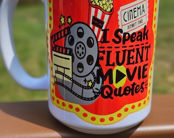 May include: White ceramic mug with a red, yellow, and black design. The design features a film reel, popcorn, a movie ticket, and the text "I Speak Fluent Movie Quotes".