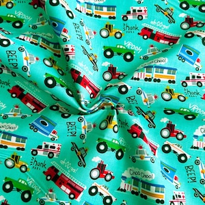 Könnte beinhalten: Ein türkisfarbener Stoff mit einem sich wiederholenden Muster aus bunten Spielzeugfahrzeugen. Die Fahrzeuge umfassen ein rotes Feuerwehrauto, ein blaues Polizeiauto, einen grünen Monstertruck, einen gelben Schulbus und einen Traktor. Der Stoff hat die Worte "Choo Choo!" und "Beep!" aufgedruckt.