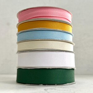 Könnte beinhalten: Ein Stapel von sieben Spulen aus Grosgrain-Band in verschiedenen Farben. Die Farben umfassen Rosa, Gold, Hellblau, Creme, Weiß und Dunkelgrün. Das Band ist ordentlich aufeinander gestapelt, wobei die Kanten der Spulen sichtbar sind.